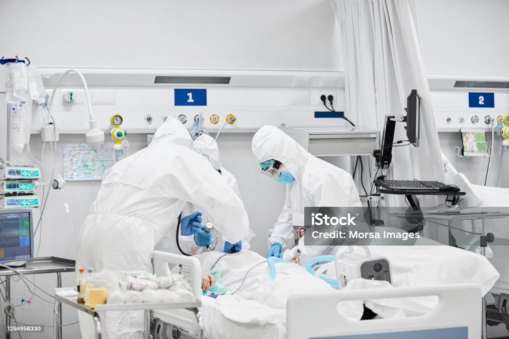 Frontline Workers Treating Male Patient in ICU Frontline workers treating male patient in ICU. Team of doctors are wearing white coveralls. They are in hospital during COVID-19. Coronavirus Stock Photo