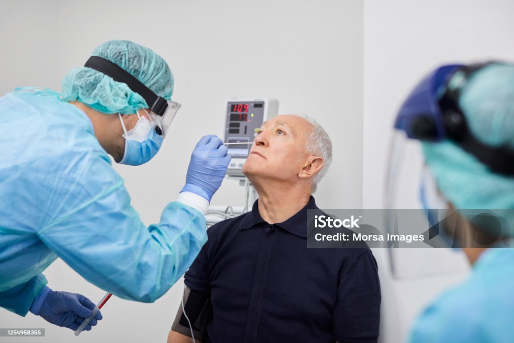 Doctor Taking Coronavirus Sample From Male's Nose, PCR. Doctor taking coronavirus sample from male patient's nose. Frontline worker is in protective workwear. They are at hospital during epidemic. Scientific Experiment Stock Photo