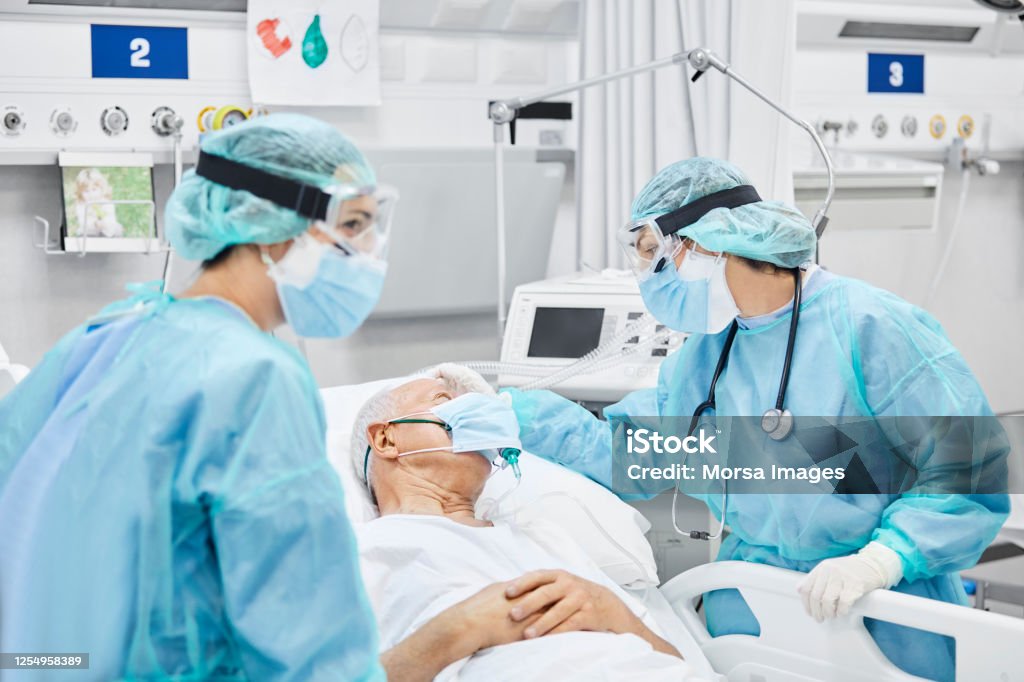 Female Doctor Talking With Patient Along Coworker Female doctor talking with patient along coworker in ICU. Man is lying on bed amidst essential workers. Healthcare workers are in protective workwear. Patient Stock Photo