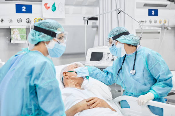 Female Doctor Talking With Patient Along Coworker Female doctor talking with patient along coworker in ICU. Man is lying on bed amidst essential workers. Healthcare workers are in protective workwear. intensive care unit stock pictures, royalty-free photos & images