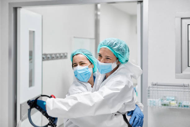 Happy Frontline Coworkers Embracing at Hospital Happy female frontline coworkers embracing each other. Healthcare workers are wearing white coveralls. They are at hospital during coronavirus. frontline worker mask stock pictures, royalty-free photos & images