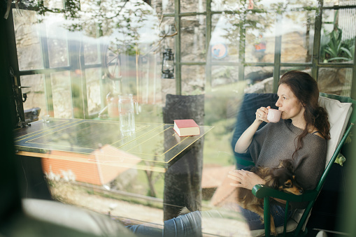 Beautiful woman is sitting in swinging chair on glass terrace with adorable dog in arms and enjoys a cup of coffee while watching nature trough the window of a glazed living room