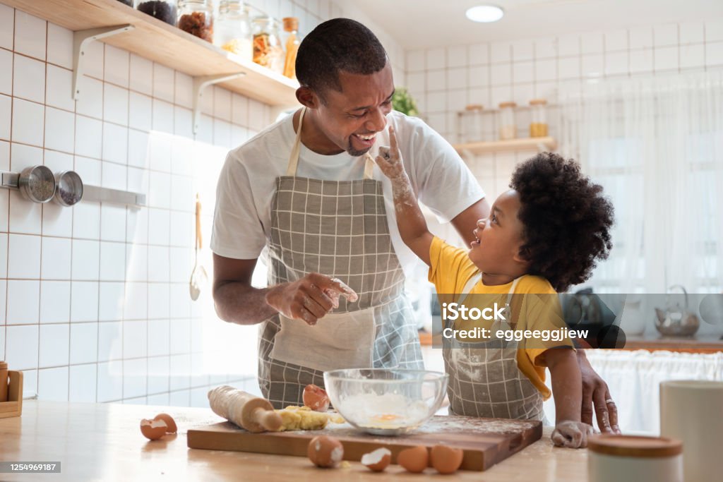 African Father and son enjoying during bake cookies at home together. African family have fun cooking baking cake or cookie in the kitchen together, Happy smiling Black son enjoy playing and touching his father nose with finger and flour while doing bakery at home. Family Stock Photo