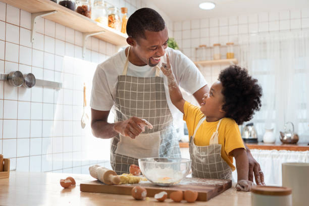 African Father and son enjoying during bake cookies at home together. African family have fun cooking baking cake or cookie in the kitchen together, Happy smiling Black son enjoy playing and touching his father nose with finger and flour while doing bakery at home. apron photos stock pictures, royalty-free photos & images