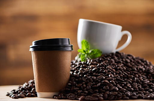 Closeup shot of a paper cup on a surface, with a teacup and mint leaf resting on a pile of coffee beans in the background