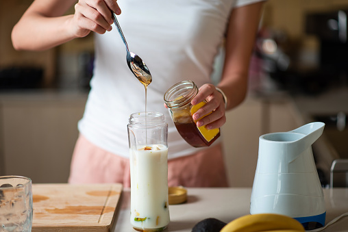 Woman adding honey while making avocado and banana smoothie for a juice with healthy breakfast at home