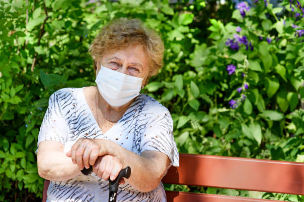 An elderly woman in a protective medical N95 Face Mask  is sitting on a park bench. Portrait of a pretty old woman with a stick in her hands, outdoors. senior adult women park bench 70s stock pictures, royalty-free photos & images