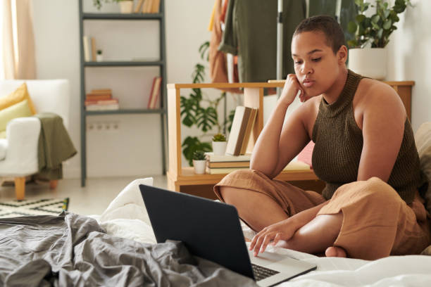 istock Serious young woman with buzz cut sitting on bed and leaning head on hand while surfing net on laptop 1254979634