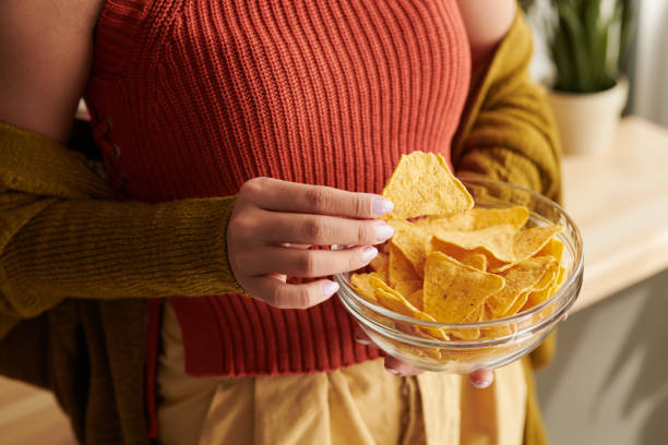 plan rapproché de la femme méconnaissable dans le dessus lumineux et le cardigan mangeant des nachos de bol à la maison, concept malsain de nourriture - tortilla chip photos et images de collection