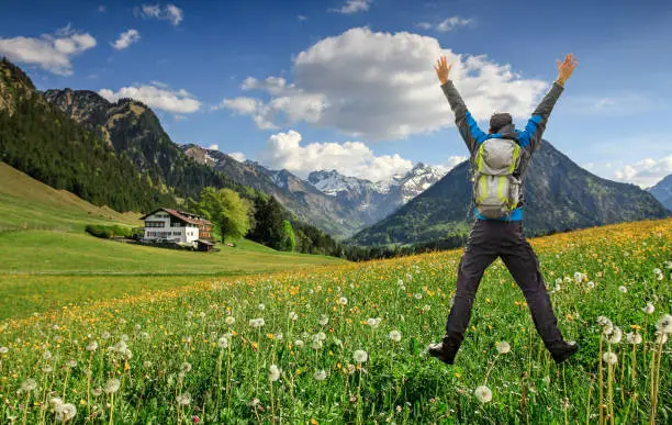 Man with Backpack and hiking equipment jumps with raised arms on beautiful dandelion flower meadow. Snow-covered mountains with clouds and a cottage in the background. Bavaria, Alps, Allgäu, Oberstdorf, Germany.