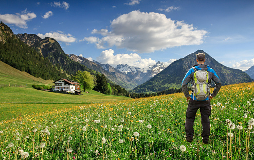 Man with Backpack and hiking equipment stands on beautiful flower meadow. Snow-covered mountains with clouds and a cottage in the background. Bavaria, Alps, Allgau, Oberstdorf, Germany.