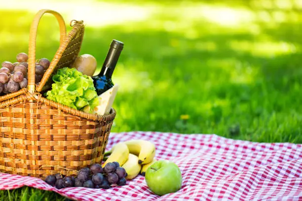 Wicker picnic basket with cheese and wine on red checkered table cloth on green grass outside in summer park, no people