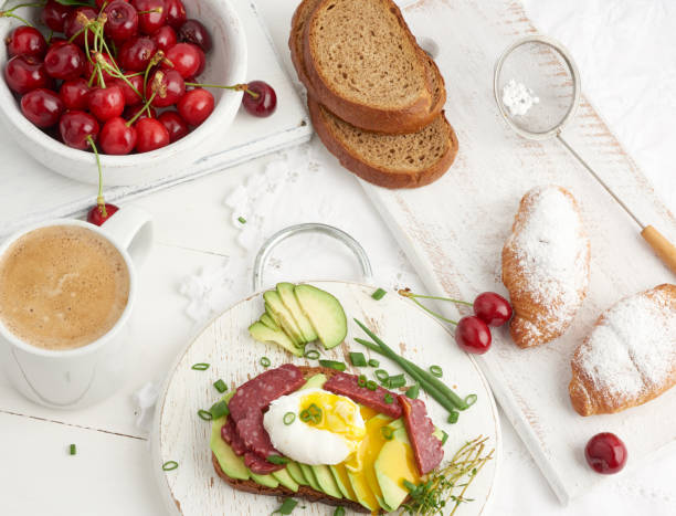 toast with poached egg and avocado on a round board, next to croissants and ripe red cherries, morning breakfast