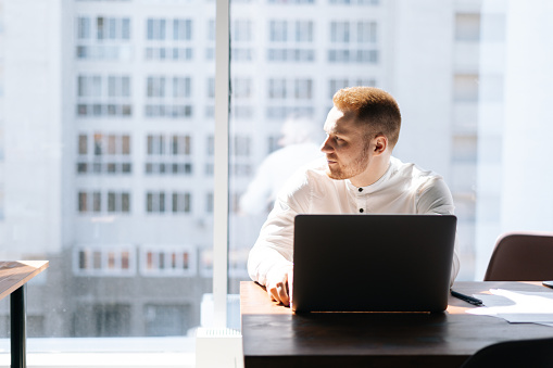 Young business man sitting at the table with laptop in the office room and looking at the side, background of large window. Concept of office working.