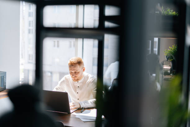 istock Attractive young business man typing on laptop doing research against window. 1254986452