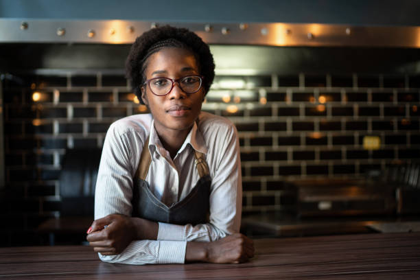 ritratto di un barista serio in piedi dietro il bancone - owner small business restaurant african ethnicity foto e immagini stock