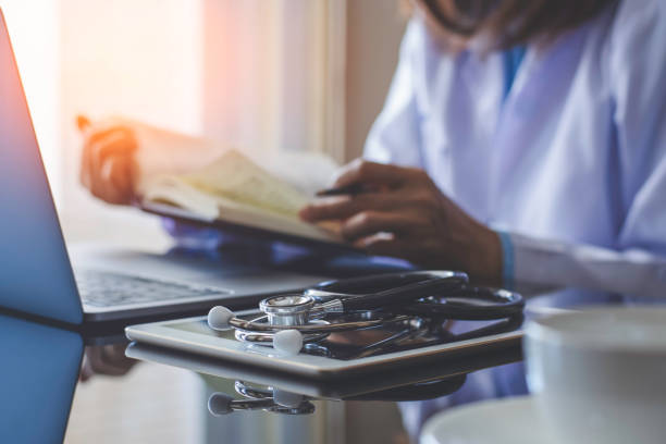 Female doctor Female doctor in white lab coat reading text book, work on laptop computer with digital tablet and stethoscope on the desk in medical room at clinic. Medical knowledge and online education concept. ipad calculator stock pictures, royalty-free photos & images