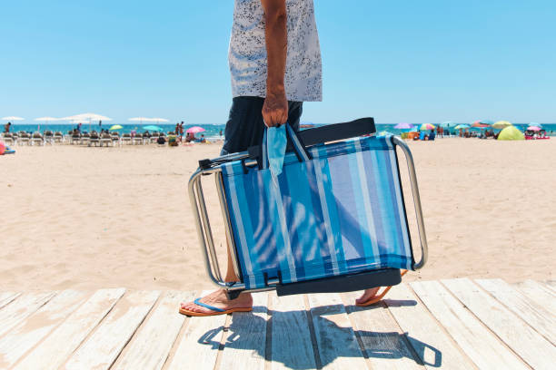 man carrying a surgical mask and a deck chair closeup of a caucasian man, wearing shorts and flip-flops, walking by a boardwalk carrying a surgical mask and a folded beach chair in his way from or to the beach, in the background foldable stock pictures, royalty-free photos & images