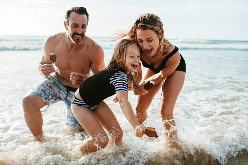 Family holding ice-cream candies and playing in sea water. Father, mother and daughter enjoying a summer weekend on the beach.