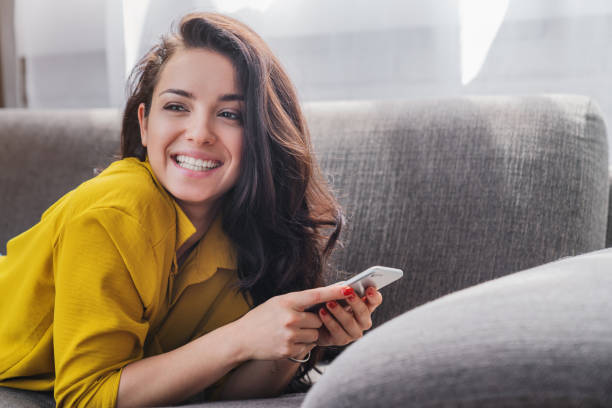 istock Young woman smiling and using mobile phone while lying on sofa at living room with look in camera 1254994111