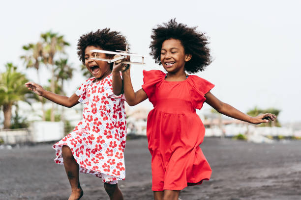 sorelle gemelle afro che corrono sulla spiaggia mentre giocano con l'aereo giocattolo in legno - stile di vita giovanile e concetto di viaggio - focus principale sul viso giusto per bambini - family american culture african culture black foto e immagini stock
