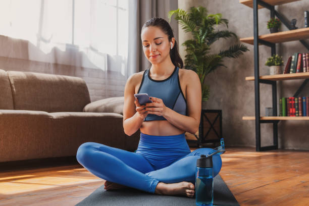 hermosa mujer con auriculares mirando el teléfono inteligente, escuchando música y relajándose después del entrenamiento - attractive female only young women music living room fotografías e imágenes de stock