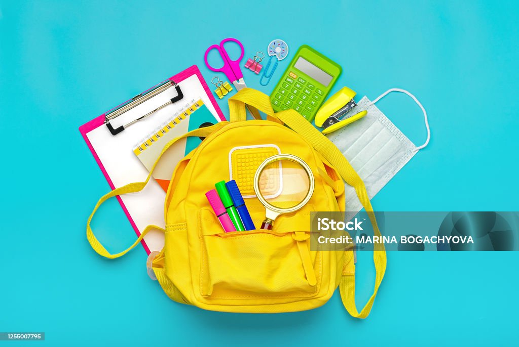 Back to school, education concept Yellow backpack with school supplies, protective medical mask, calculator, scissors isolated on blue background. Top view Copy space Flat lay Back to school, education concept Yellow backpack with school supplies, protective medical mask, calculator, scissors isolated on blue background. Top view Copy space Flat lay. Back to School Stock Photo