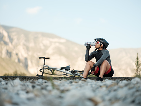 A latin cyclist woman sitting on the road next to her bike and drinking water from a water bottle.