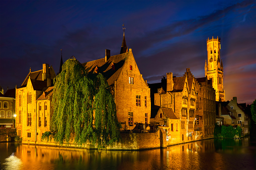 Famous view of Bruges tourist landmark attraction - Rozenhoedkaai canal with Belfry and old houses along canal with tree in the night. Brugge, Belgium