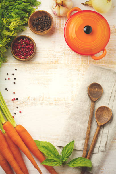 Black and red pepper, basil leaves, carrots,  ceramic pan,  wooden stand, simple old spoons and  linen napkin on  light background. Kitchen accessories concept. Selective focus. Top view. Toned image.