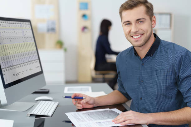 Happy successful young businessman at his desk Happy successful young businessman at his desk in the office working on a report looking at the camera with a warm smile financial advisor photos stock pictures, royalty-free photos & images