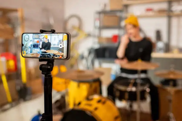 Young contemporary man in front of drumset on smartphone screen during making video about drum music for his online subscribers