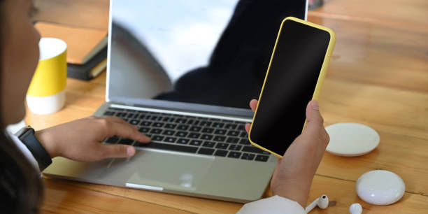 istock Cropped image of the hand is using a smartphone while sitting in front of an empty screen computer laptop. 1255012712