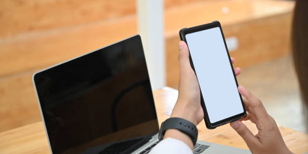 istock Cropped image woman is holding a white blank screen smartphone while sitting in front of a computer laptop. 1255012879