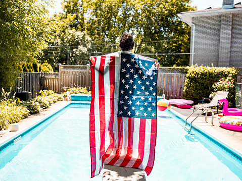 Black boy holding American National flag while standing on a diving board by the backyard pool.