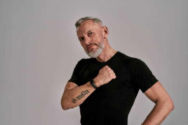 Middle aged muscular man in black t shirt looking at camera, beating his chest while posing in studio over grey background. Sport, healthy lifestyle concept
