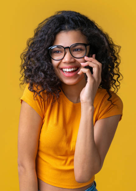 vertical shot of cute smiling casually dressed girl talking on her mobile phone isolated on bright colored yellow background - smiling casino human hand beautiful imagens e fotografias de stock