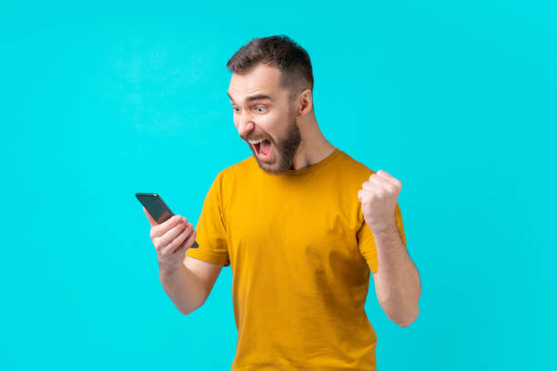 Football fan watching play live broadcast online from his mobile phone and cheering for favourite team Attractive euphoric bearded man clenching his fist in triumph. Football fan watching play live broadcast online from his mobile phone and cheering for favourite team. mobile casino games stock pictures, royalty-free photos & images