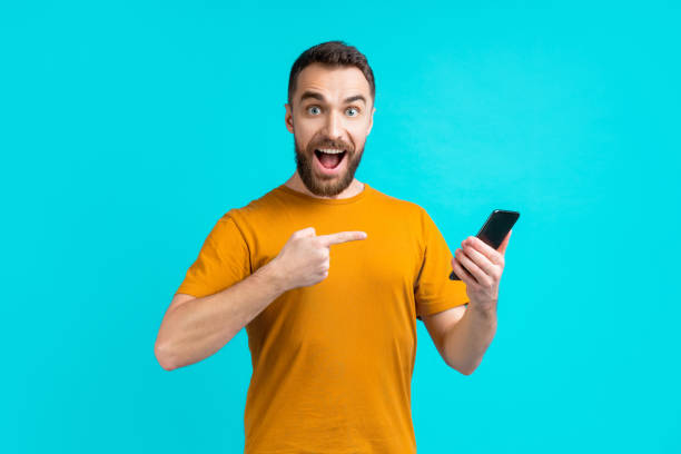 istock Excited smiling bearded man pointing at his smartphone touchscreen with index finger surprised with great news he just got with a message 1255030913