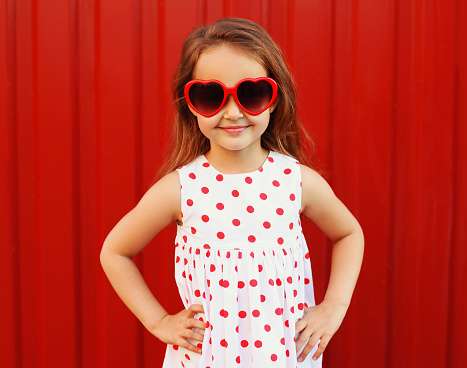 Portrait happy smiling little girl child wearing a white dress and red heart shaped sunglasses on background