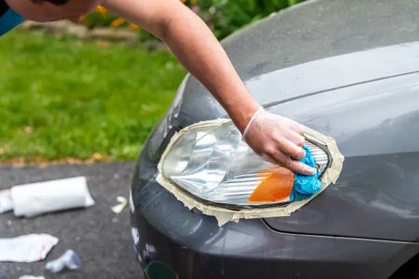 Man person worker cleaning car headlights taped to protect paint rubbing with blue shop towel closeup on driveway of house outside