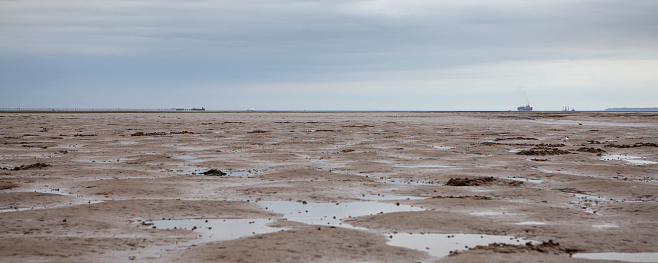 Wide panoramic view of the Thames estuary at low tide looking east towards Southend-on-Sea with pier and container ship on the horizon