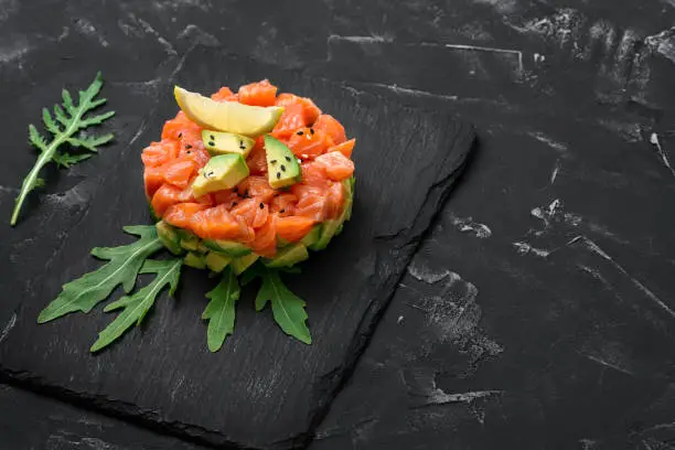 Raw salmon tartare. Fresh salmon, avocado and arugula salad in a culinary ring on a slate plate, black stone background. Selective focus