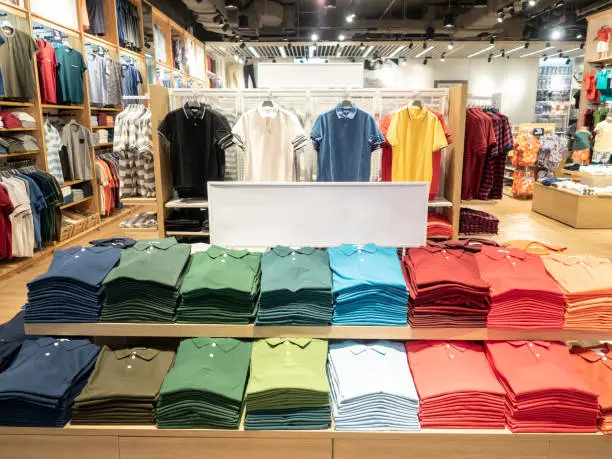 Photo of Colorful polo t shirt stack on shelf in large department store shop. Copy space on white sign board.