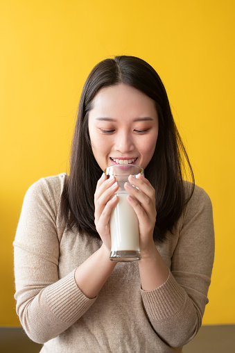 Young attractive Asian woman with closed eyes drinking milk against yellow background