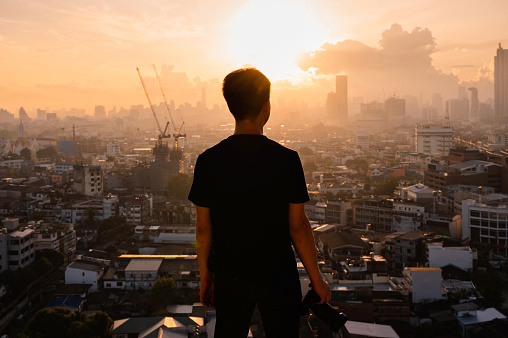 Young asian man with camera standing on top of building and view of sunrise on Bangkok city in Thailand