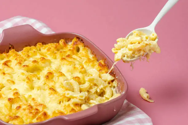 Grabbing a spoon of cheesy macaroni from the tray with mac and cheese freshly baked. Mac and cheese oven cooked in white sauce, with parmesan, mozzarella, gruyere, emmentaler and cream.