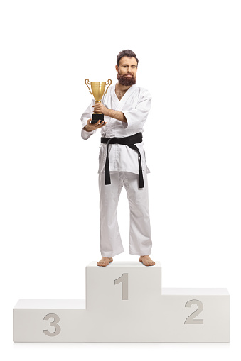 Full length portrait of a karate champion in kimono with a trophy cup on a winners podium isolated on white background