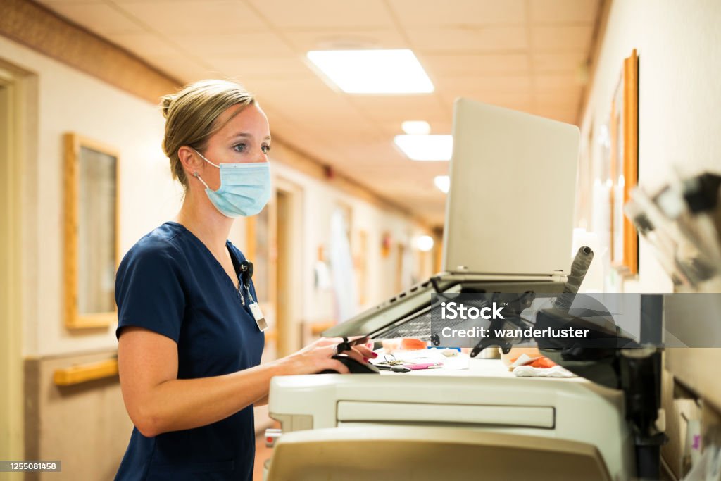 Nurse documenting during Covid-19 Registered nurse wearing a face mask documents medicine provided to a patient into a laptop computer at the nursing station at work, Indiana, USA Nurse Stock Photo