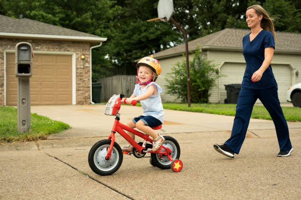 Learning how to ride a bike A nurse wearing scrubs watches her little  daugher ride her bike with training wheels before work, Indiana, USA day in the life stock pictures, royalty-free photos & images